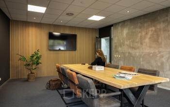 Modern conference room at Lintelerweg 40, Hengelo, with a large wooden table, chairs, TV screen, and decorative plant. A person is working on a laptop.