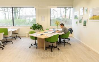 Modern office space rental in Herentals at Atealaan 34A, featuring large windows, green chairs, and a few tables with one person working on a laptop.