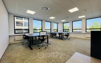Bright office space in Hilversum featuring multiple black desks and mesh chairs by large windows with a view, available for rent.