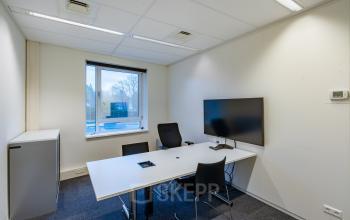 Modern office space rental at Olympia 4 in Hilversum features a sleek meeting room with a large table, chairs, and wall-mounted screen. Natural light floods through a window, enhancing productivity.