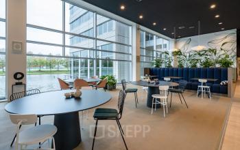 Modern workspace at Polarisavenue 1, Hoofddorp, featuring tables, chairs, a bench, plants, large windows, and outside view of building and water.