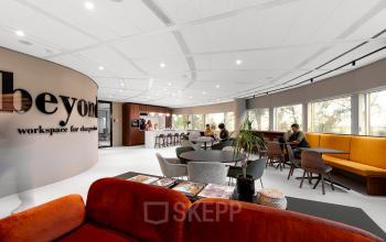 Modern furnished office space featuring individuals working and interacting in an open area with large windows and comfortable seating.