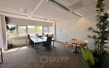 A well-lit office space available for rent at Planetenweg 5, Hoofddorp. The room features a spacious table with chairs, a small round meeting table, and large windows with natural light.