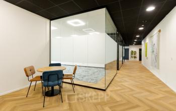 Modern office space with glass walls and wood flooring, featuring a round table with chairs, located at Industriepark 5B, ideal for office space rental.