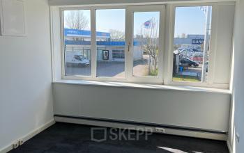 An office space at Zwettestraat 15 in Leeuwarden featuring large windows with views of the street and surrounding buildings, ideal for rent. The room is unfurnished, showcasing a clean slate for business needs.