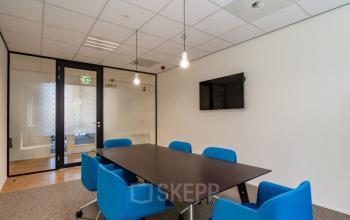 Conference room at Schipholweg 55-89, Leiden, featuring a rectangular table with blue chairs, glass walls, a mounted TV, and modern lighting fixtures.