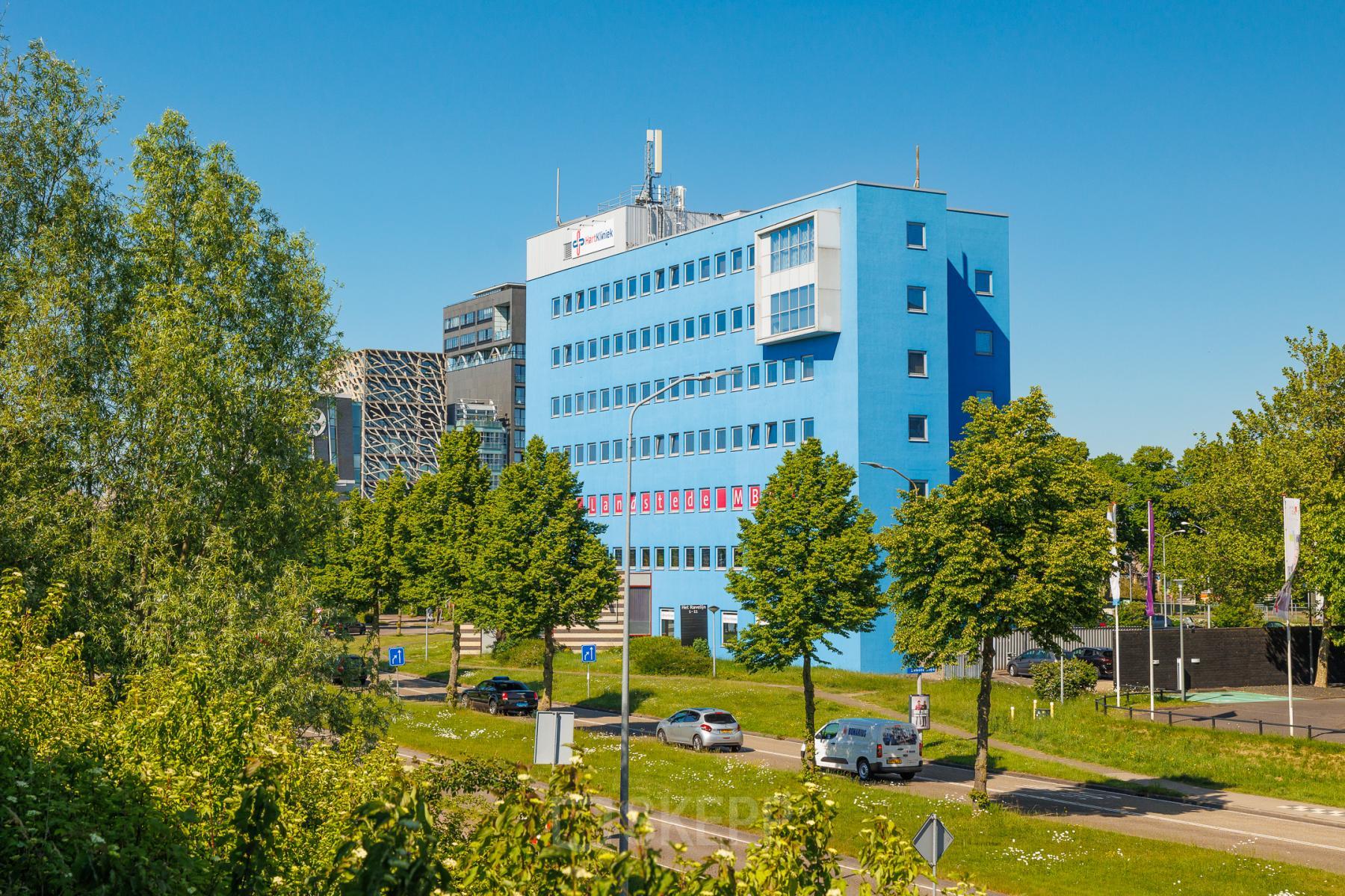 Exterior view of a blue office building at Het Ravelijn 1-11, Lelystad, surrounded by trees and parking. Ideal location for office space rental with ample natural light.