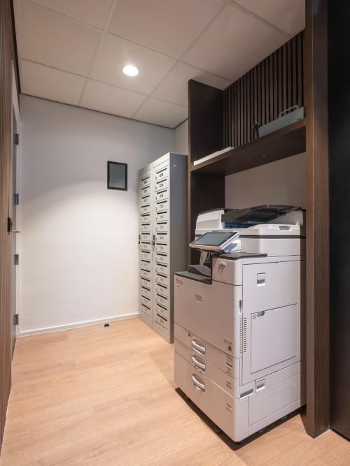 Office interior with wood flooring, featuring a large multifunction printer and mail slots, ideal for efficient office tasks. Located in Maastricht-Airport, this space offers practical amenities for office space rental.
