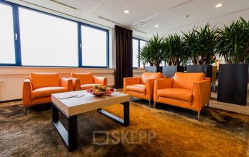Office lounge area at Europalaan 24, Maastricht-Airport, with four orange chairs, a wooden table, and potted plants near the windows.
