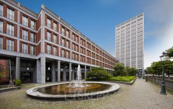 Contemporary office space rental at Stationsplein 8K, Maastricht, with a fountain in the foreground and a clear sky above the modern facade.