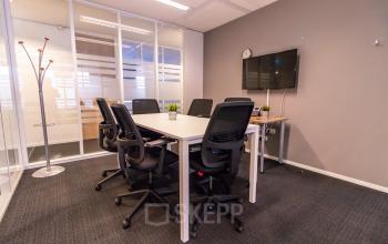 A furnished conference room at Robert Schumandomein 2 in Maastricht features a table with six chairs, a wall-mounted screen, and a minimalistic design, perfect for office space rental.