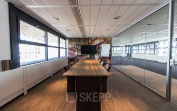 Modern conference room at Boschstraat 21, Maastricht with a long wooden table, black chairs, large windows, glass walls, and a wall-mounted TV.