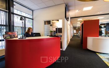 Office space rental at Boschstraat 21, Maastricht, featuring a modern reception area with a curved red desk, adjacent workstations, and large windows.