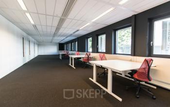 Office space for rent at Boschstraat 21, Maastricht, featuring a row of desks with chairs, large windows, and ample lighting.