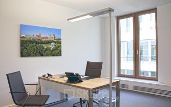 Modern office space at Theresienstraße 1, Munich Maxvorstadt, featuring a well-lit room with two chairs, a desk, office equipment, and a large window with a cityscape view.