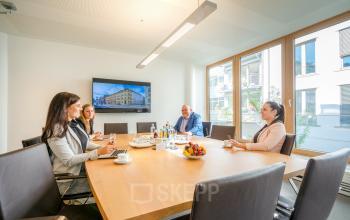 Meeting room with a wooden table and four people in business attire having a discussion at Theresienstraße 1, Munich Maxvorstadt. Large windows provide natural light. Office space rental available.