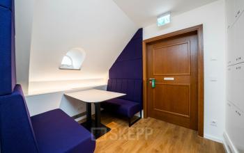 Kompakter Büroraum in Ludwigstraße 8, München Maxvorstadt mit lila Sitzbank, Tisch und Holztür. Ideal für informelle Besprechungen oder Pausen. Bürofläche mieten in zentraler Lage.