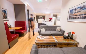 Moderner, gut möblierter Büroraum mit gemütlichen Sitzgelegenheiten zur Miete in München Maxvorstadt, Hopfenstraße 8. Personen arbeiten in einem ansprechenden, offenen Arbeitsbereich.