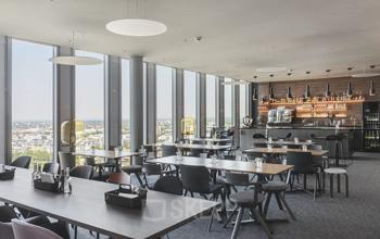 Helles Büro mit Panoramafenstern in der Mies-van-der-Rohe-Straße 8, München Schwabing, mit modernem Mobiliar und einer Bar im Hintergrund. Optimal für Büroraum mieten.