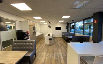 Modern office kitchen and lounge area with tables, chairs, booths, and a large screen on the wall at Rijksweg 79, Naarden. Suitable for office space rental.