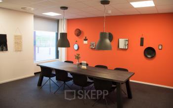 Modern office space with a long wooden table, black chairs, and orange accent wall at Rijksweg 79, Naarden. Suitable for meeting or collaborative work.