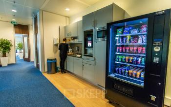 A furnished office space rental in Nijmegen featuring a pantry area with a person preparing a drink beside a stocked vending machine.