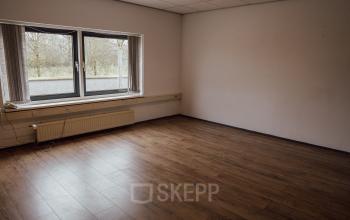 An empty interior space with wooden flooring and a large window, ideal for those seeking office space rental in Oosterhout Nb.