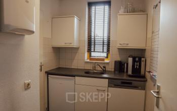 Compact kitchenette with white cabinets, small fridge, coffee machine, and a window with blinds in an office space to rent.
