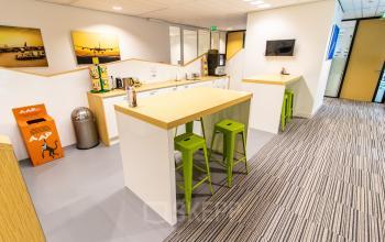 Brightly lit office space at Rotterdam Airportplein 22 featuring a kitchenette with green stools and a coffee machine, ideal for office space rental.