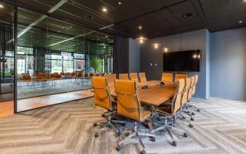 A modern conference room with a long wooden table and brown swivel chairs, equipped with video conferencing facilities. The glass wall reveals additional seating.