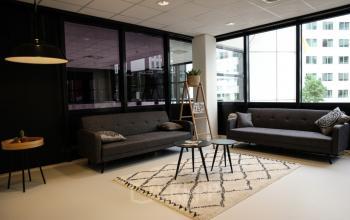 Modern lounge area with grey sofas, a patterned rug, and small tables in an office building at Marten Meesweg 8-10, Rotterdam Alexander. Large windows provide natural light.