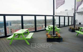 Rooftop terrace with green picnic tables, planters, and a view of surrounding buildings in Rotterdam Alexander, ideal for a break from the office space rental at Hoofdweg 66.