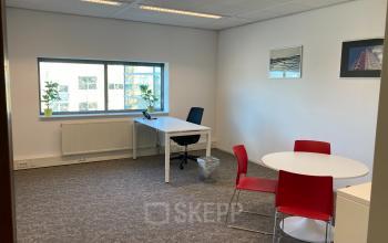 Bright office space at Hoofdweg 66, Rotterdam Alexander, featuring a desk by the window, round table with red chairs, and white walls with framed art.