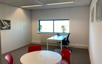 Office space with modern furnishings located at Hoofdweg 66 in Rotterdam Alexander; features a large window, desk with chair, round table with red chairs, and framed artwork.