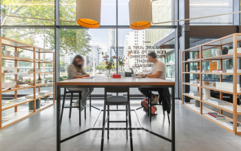 Modern workspace at Rotterdam Central Station featuring large windows, natural light, and individuals collaborating at a high table with shelving units around.