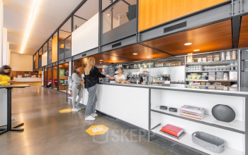 Modern office pantry at Hofplein 20, Rotterdam Central Station, featuring shelves with kitchenware, a coffee station, and people engaging in discussions. Ideal for rent office space enthusiasts.