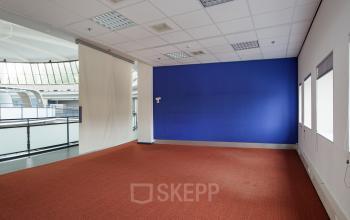 Office space for rent at Kruisplein 15-25, Rotterdam Central Station, featuring a bright room with orange carpet and blue accent wall, large windows, and ample lighting.