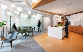 Modern office space at Stationsplein 45, Rotterdam Central Station, featuring a well-lit lounge area with tables, chairs, plants, and a minimalist design. People are seen engaging in business activities.