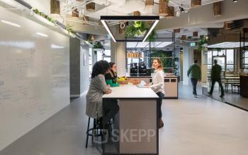 Modern office lounge area with a high table and stools, whiteboard wall, and plants hanging from the ceiling at Stationsplein 45, Rotterdam Central Station.