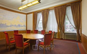 A classic conference room with a round table and eight red upholstered chairs at Schiekade 830 in Rotterdam Center. Ideal for office space rental, the room features large windows and elegant drapery.