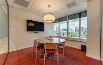 Cozy meeting room at Blaak 555, Rotterdam Center, featuring a round table, modern chairs, a large wall-mounted screen, and a window view of the cityscape. Perfect for office space rental needs.