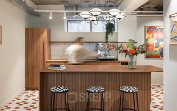Stylish office lounge area with modern wooden furniture, art on the walls, and a person making coffee at the counter.