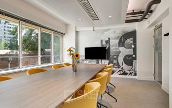 Bright conference room with a long table and yellow chairs, featuring large windows and a modern wall design. Ideal office space rental in Rotterdam Center at Coolsingel 104.