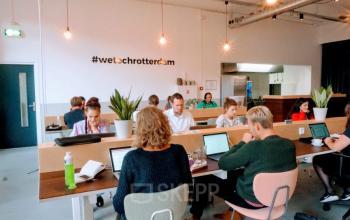 Open workspace with people working at shared desks, featuring modern lighting and indoor plants at Schiedamse Vest 154, Rotterdam Center, Rotterdam. Ideal office space rental for professionals.