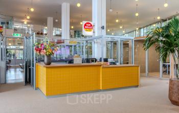 Spacious and bright office to rent at Van Nelleweg 1, Rotterdam Delfshaven, featuring a welcoming yellow-tiled reception area with glass partitions and decorative plants.