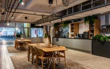 A modern office space at Van Nelleweg 1, Rotterdam Delfshaven, featuring wooden tables, chairs, potted plants, and a kitchenette, ideal for office space rental.