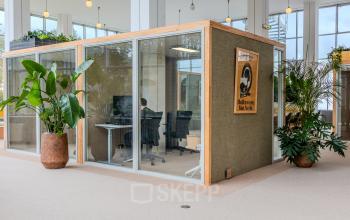 Office space rental at Van Nelleweg 1, Rotterdam Delfshaven. A person works at a desk inside a glass-walled office unit, surrounded by plants and modern furnishings.