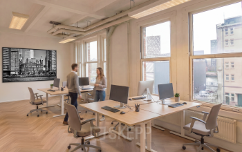 Bright office space with modern desks and computers, situated in Rotterdam Delfshaven. Two people are discussing at a desk. Ideal for office space rental.