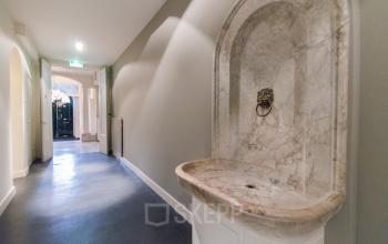Spacious corridor in a Rotterdam office with a classical marble basin, leading towards an open entrance, ideal for office space rental.