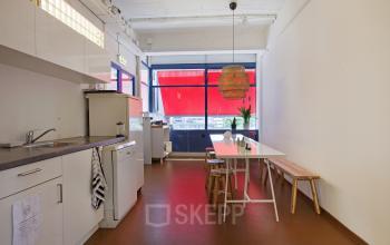 Pantry area in the office space rental at Vijverhofstraat 47, Rotterdam North, featuring a kitchenette, a white table, wooden benches, and bright red flooring, with large windows providing natural light.